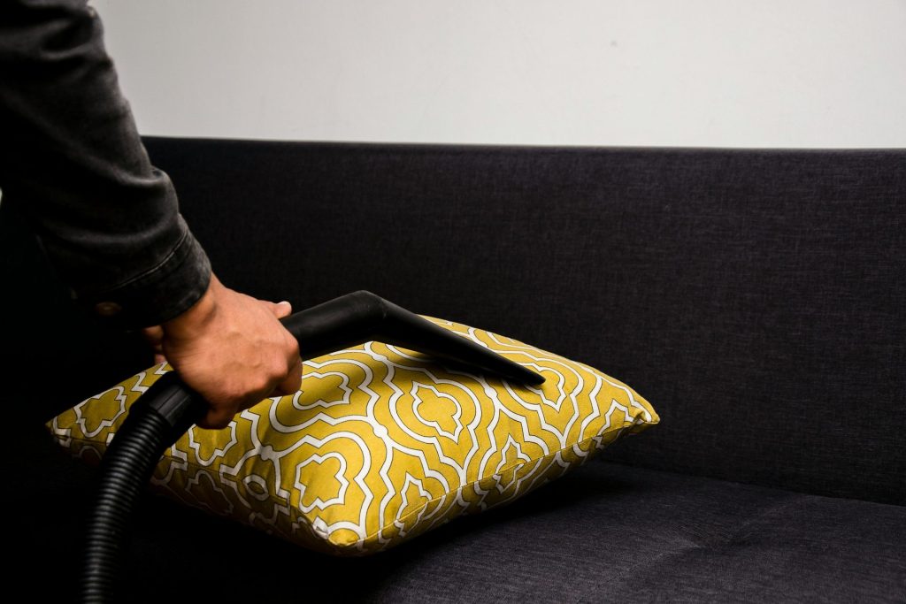 Close-up of a person vacuuming a patterned pillow on a dark sofa indoors.